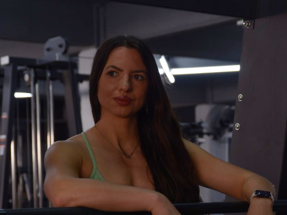 Maia Soldano en el gimnasio, apoyada sobre una barra con top deportivo verde, mirando hacia un costado con una sonrisa leve.