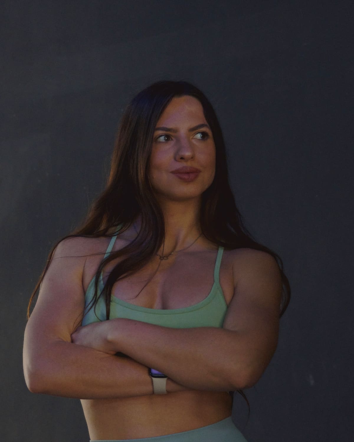Maia Soldano, nutricionista, de pie con los brazos cruzados y media sonrisa, con top deportivo verde salvia, sobre fondo oscuro.