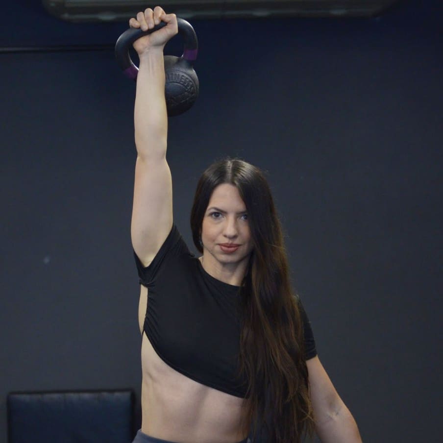Maia Soldano sosteniendo una pesa rusa con el brazo extendido sobre la cabeza, mirando a la cámara.